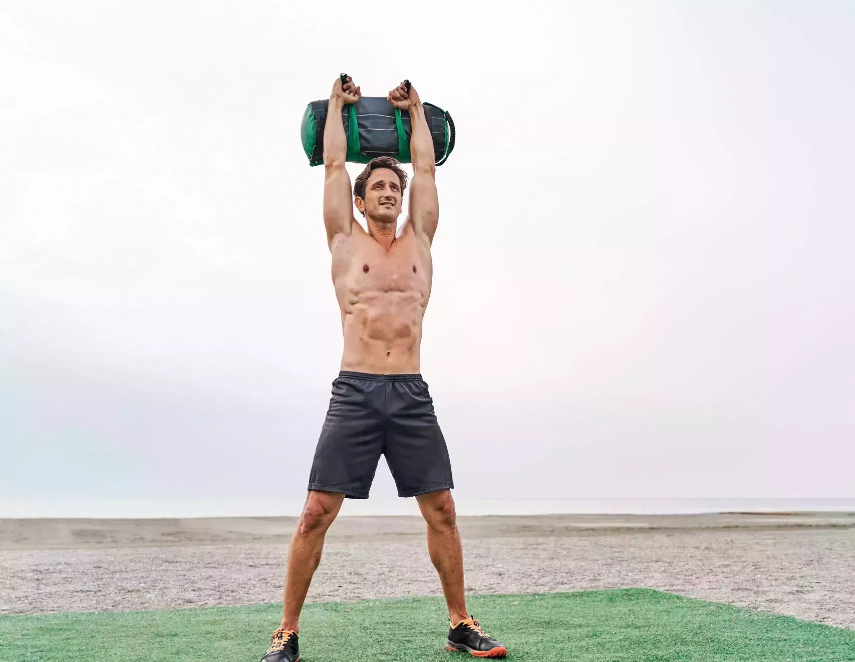 young muscular man training on the beach shirtless doing a overhead front squat lifting a sandbag