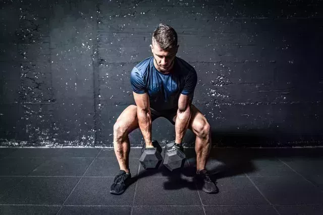 young strong fit muscular sweaty man with big muscles strength cross workout training with dumbbells weights in the gym dark image with shadows real people