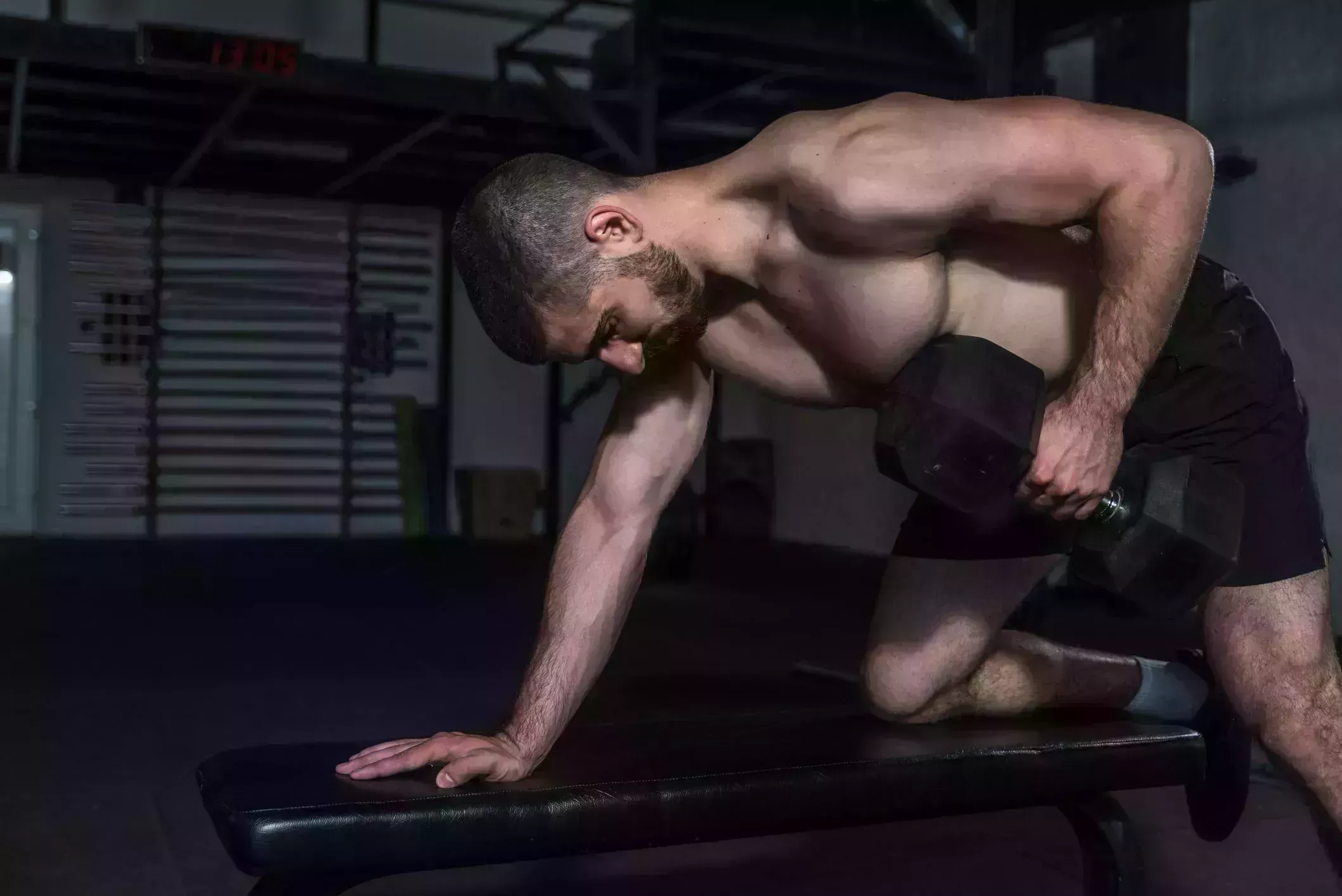 young concentrated sweaty fit muscular strong man doing hardcore workout training for back muscles with heavy dumbbell weight on the gym bench in the gym, dark image real people exercising