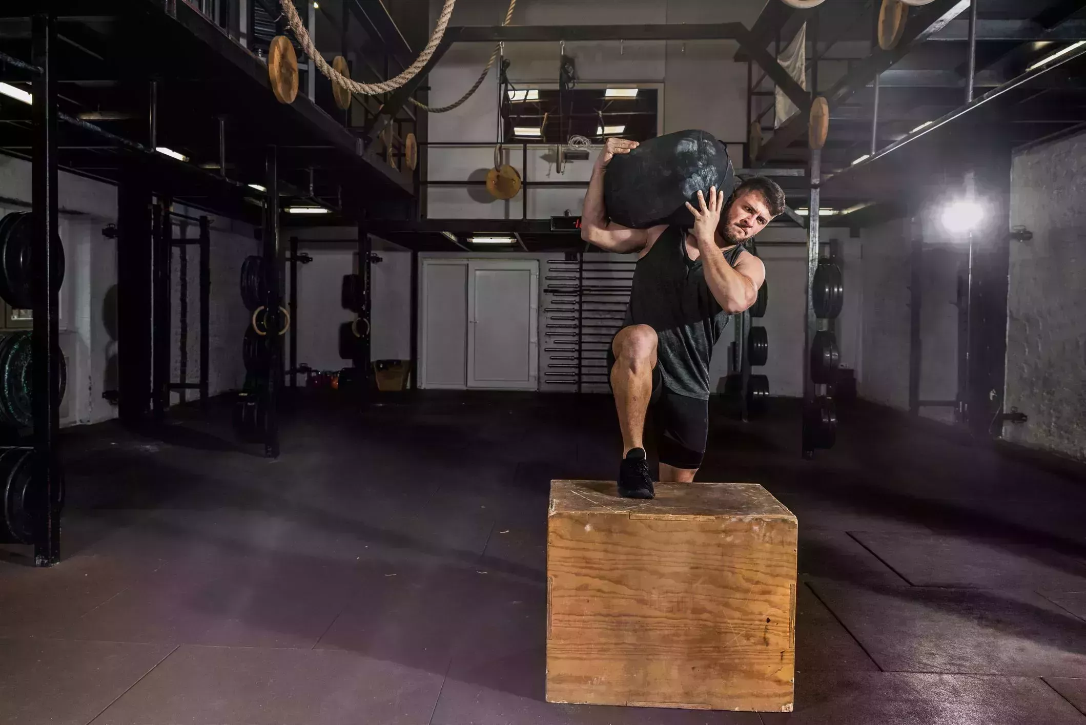 young strong sweaty fit muscular man with big muscles holding heavy sandbag on his shoulder with his hands and climb on the jump box for legs cross workout training in the gym
