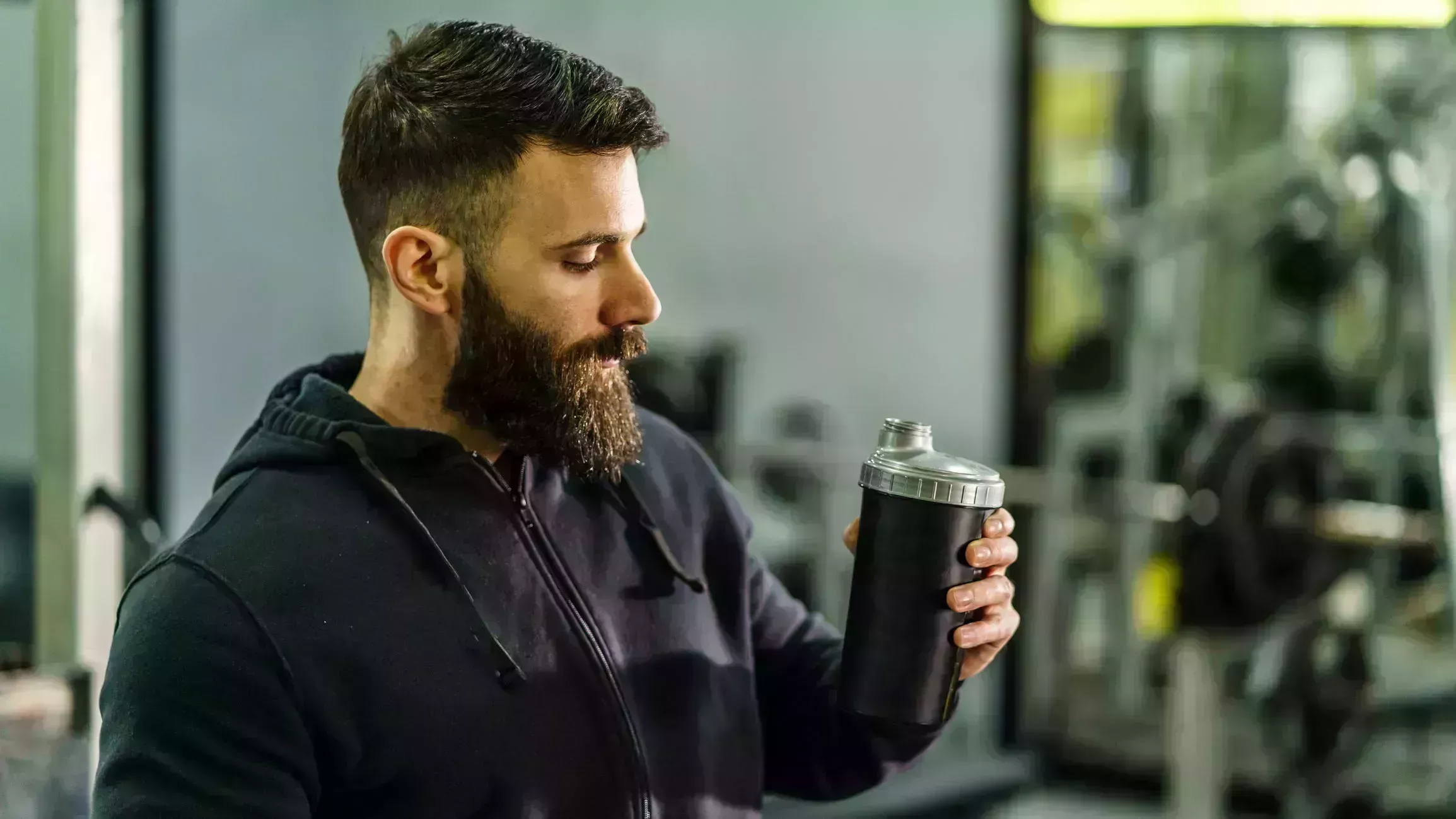 front view portrait of young caucasian man athlete in black hoodie male standing in the gym holding protein supplement shaker supplementation in training waist up black hair and beard copy space drink