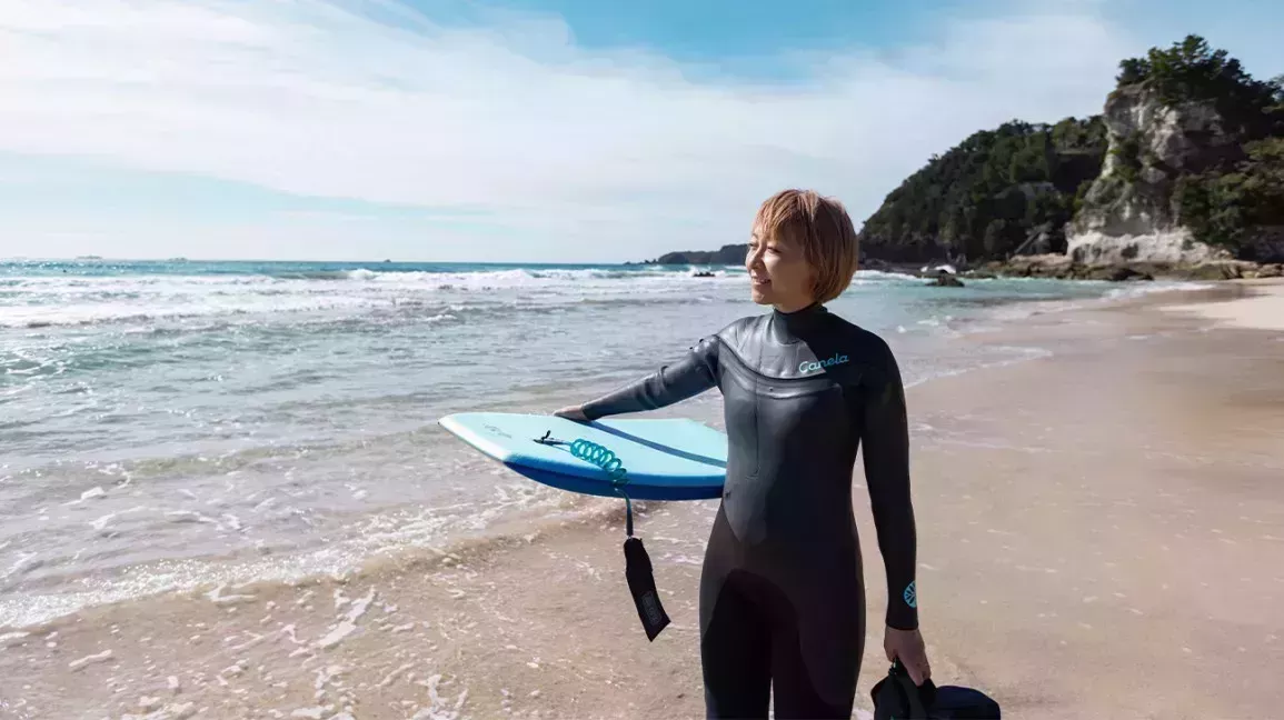Person with bobbed hair wearing wetsuit stands on beach with surfboard 1