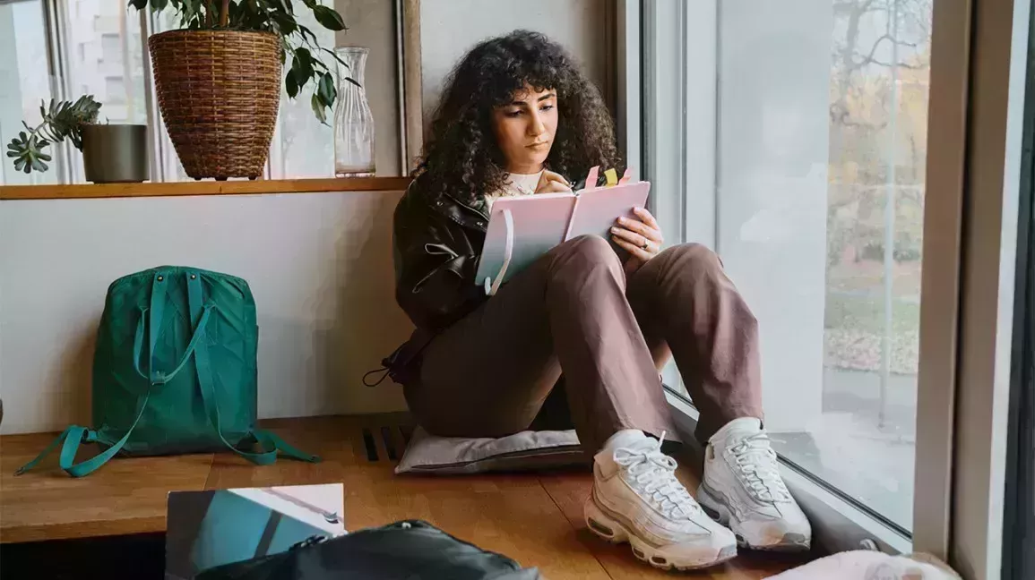 Older teenage with long curly hair sitting by window studying 1