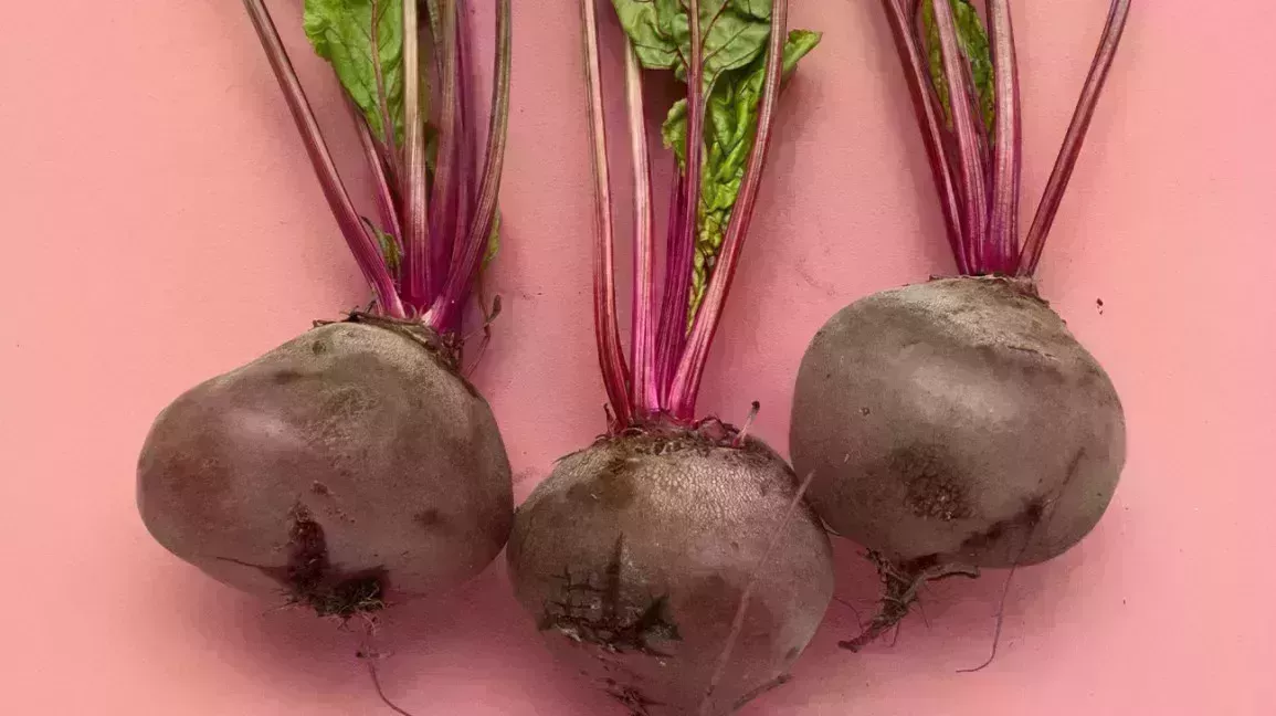 three raw beets with stems and leaves against a pink backdrop