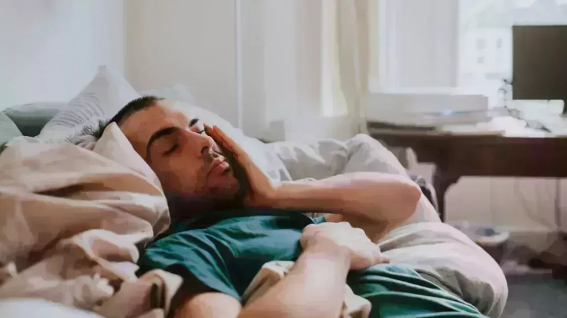 A man lies on a bed, with his eyes closed, holding the side of his face, concerned about a popped blood vessel in his eye.