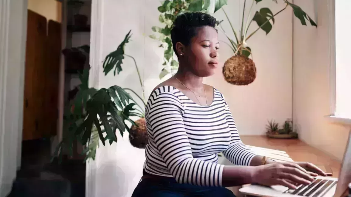 person sitting on desk, typing on laptop. Plants are behind them