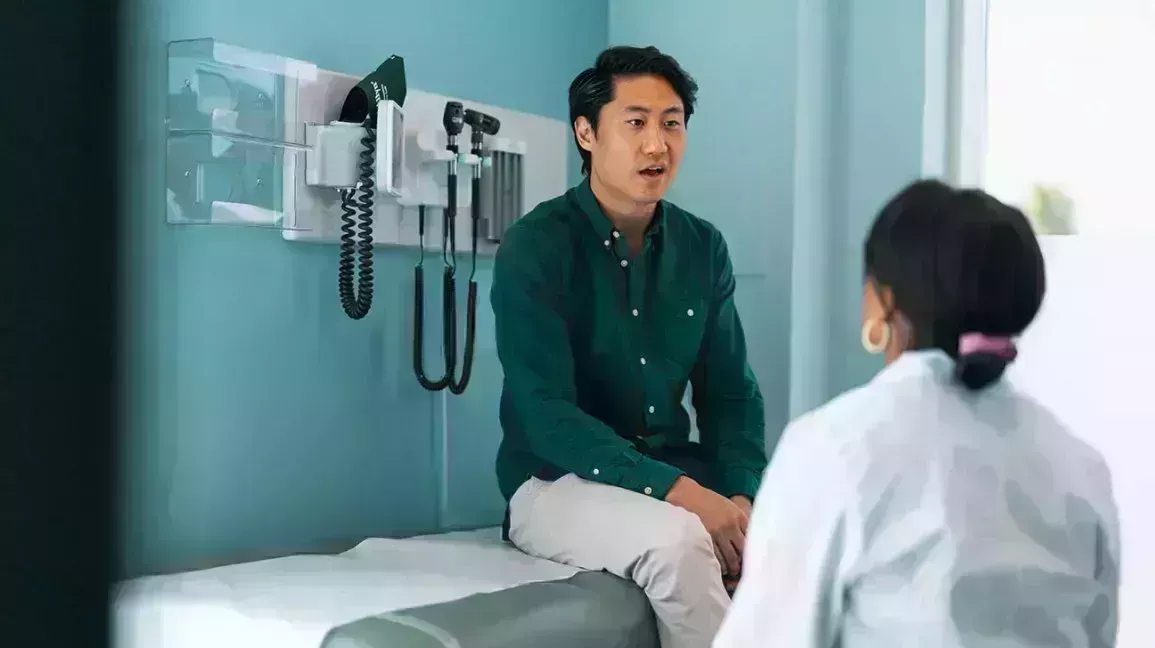 Man sitting on exam table in a doctor's office. There are doctor's tools behind him. He is wearing a dark green shirt and talking to the doctor. The doctor is standing in front of this man and she is looking up at him while he is talking.
