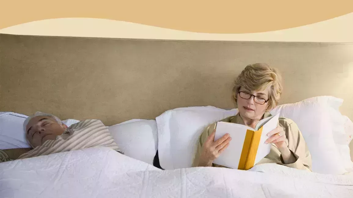One person reading in bed while another person sleeps.