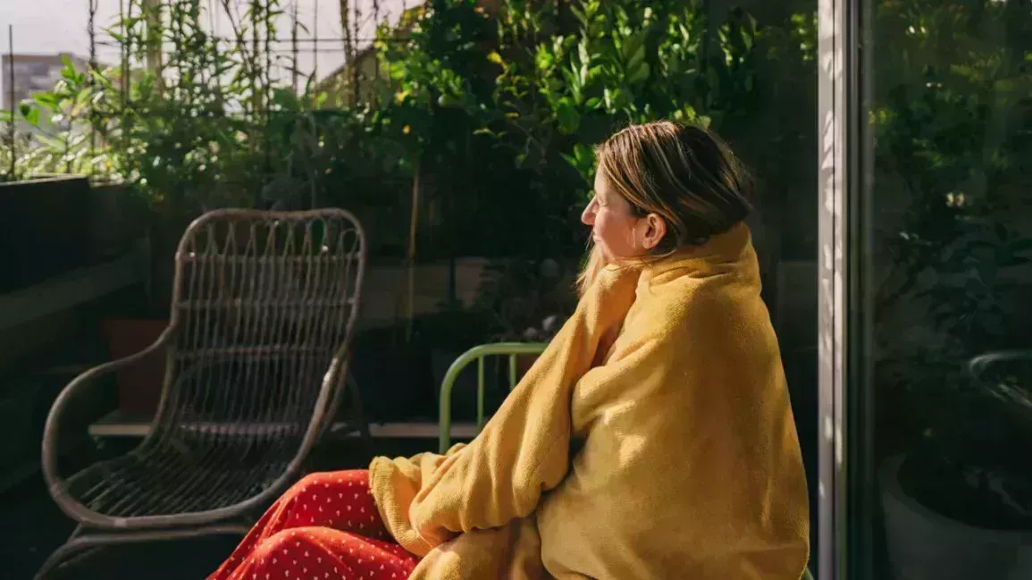 A woman sits outside with a blanket wrapped around her, trying to get some vitamin D from the sun.