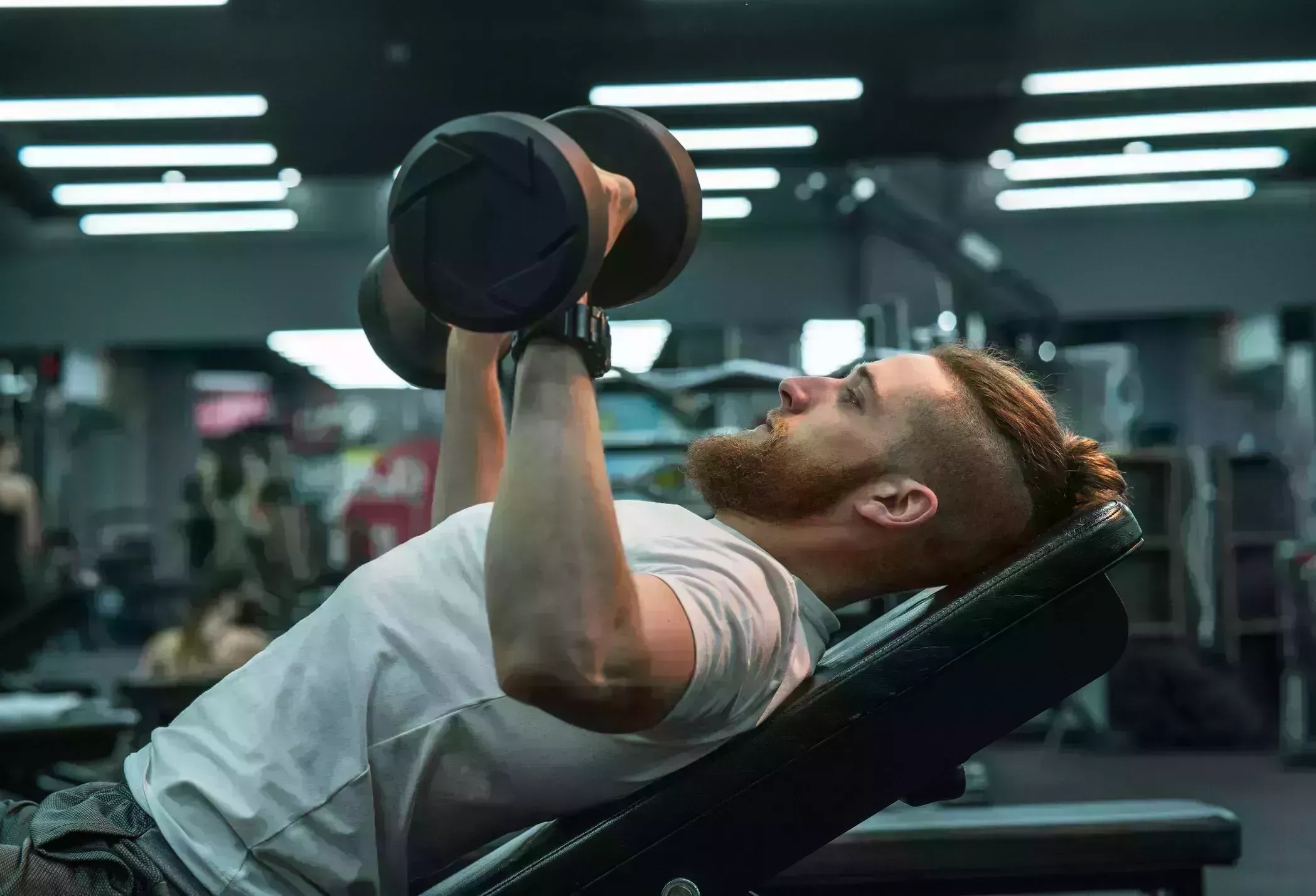 a man in sportswear lies on the gym bench and workout on his chest and arms with dumbbells